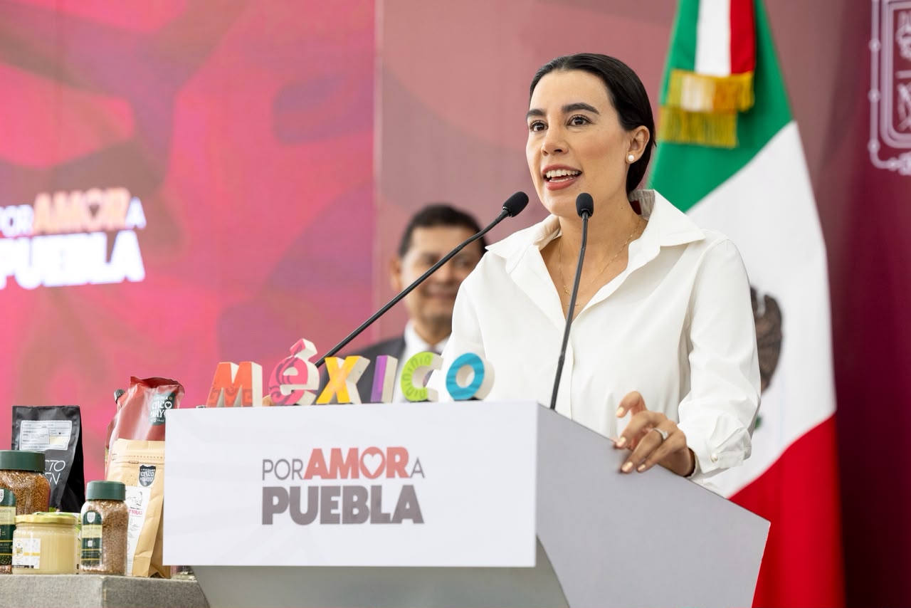 Josefina Rodríguez Zamora, Secretaria De Turismo Del Gobierno De México, Sonríe Mientras Habla Desde Un Presídium Blanco Con La Leyenda &Quot;Por Amor A Puebla&Quot; Y Letras Multicolores Que Forman La Palabra &Quot;México&Quot;. A Su Lado Se Observa La Bandera De México Y, En Primer Plano A La Izquierda, Una Exhibición De Productos Artesanales Y Gastronómicos Locales Como Café Y Miel.