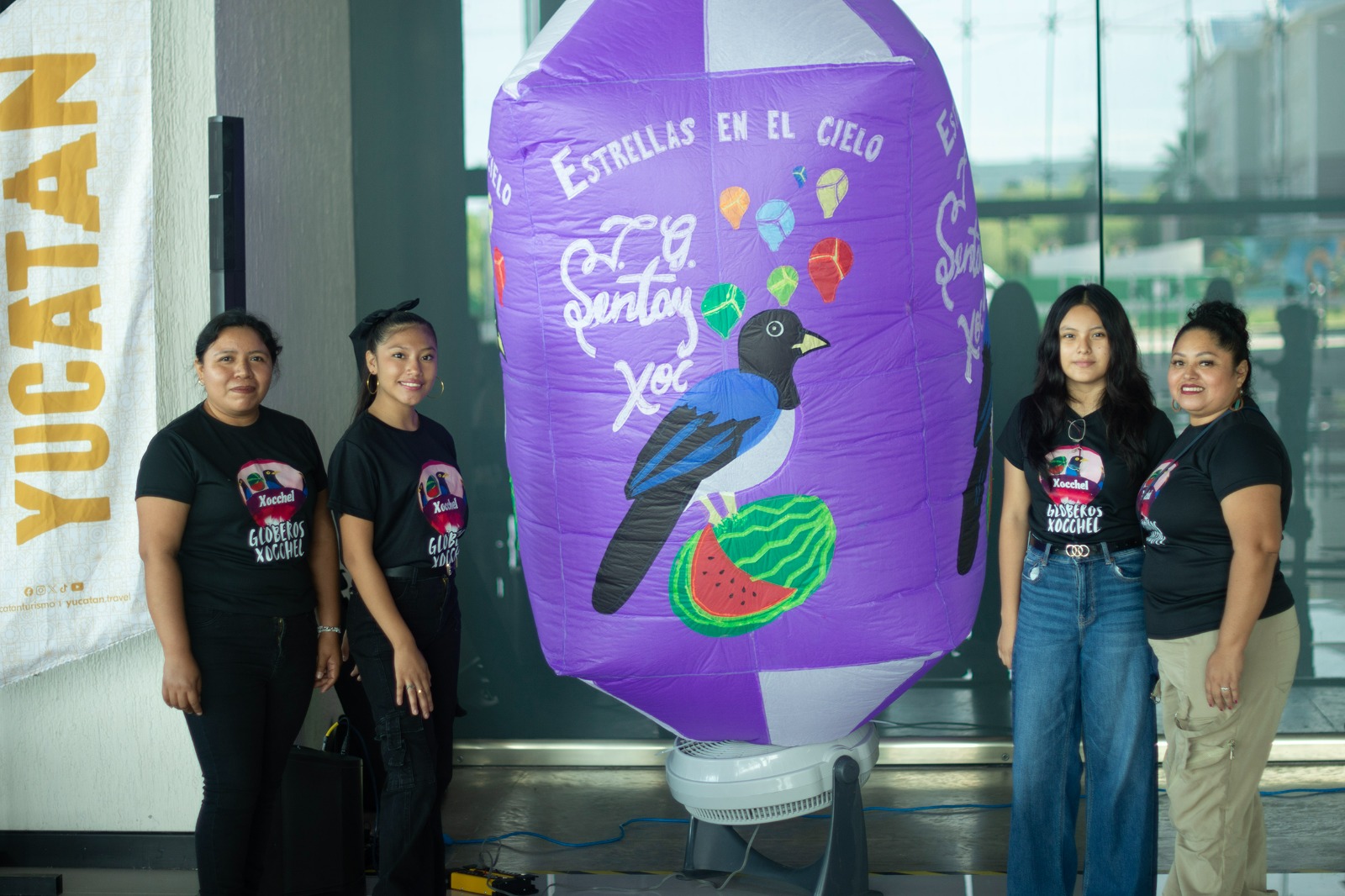 &Quot;Cuatro Mujeres Artesanas Posan A Los Lados De Un Gran Globo De Papel China Morado Inflado Con Aire, Decorado Con El Dibujo De Un Ave Sobre Una Sandía Y La Frase Estrellas En El Cielo.&Quot;