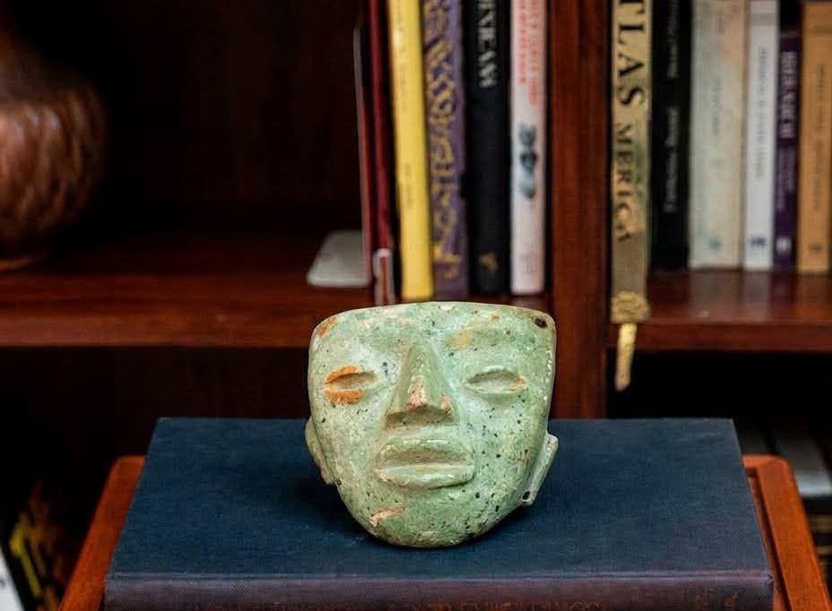 Un legado que regresa a casa: coleccionista japonés devuelve máscara prehispánica a méxico la revista del sureste mx Primer Plano De Una Máscara Prehispánica De Color Verde Jade, Con Rasgos Estilizados Y Desgastados, Colocada Sobre Un Libro Antiguo De Tapa Azul Oscuro. De Fondo, Se Aprecian Estantes De Madera Repletos De Libros Borrosos.