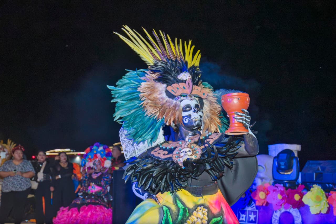 Una Niña Disfrazada Y Maquillada De Catrina Posa De Pie En Una Escalinata Decorada Con Papel Picado Y Alebrijes, Luciendo Un Vestido Tradicional Mexicano De Color Fucsia Y Un Tocado De Flores.