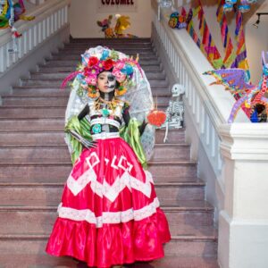 Una Niña Disfrazada Y Maquillada De Catrina Posa De Pie En Una Escalinata Decorada Con Papel Picado Y Alebrijes, Luciendo Un Vestido Tradicional Mexicano De Color Fucsia Y Un Tocado De Flores.