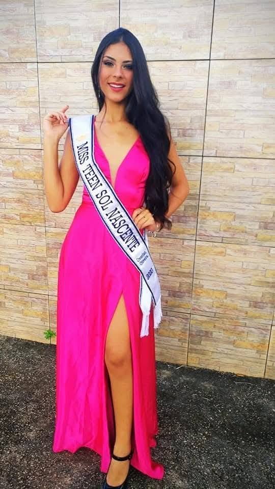 Kamila Rodrigues Sonríe De Pie Vistiendo Un Vestido Largo Fucsia Con Abertura Y Una Banda De Certamen De Belleza Cruzada Al Pecho.