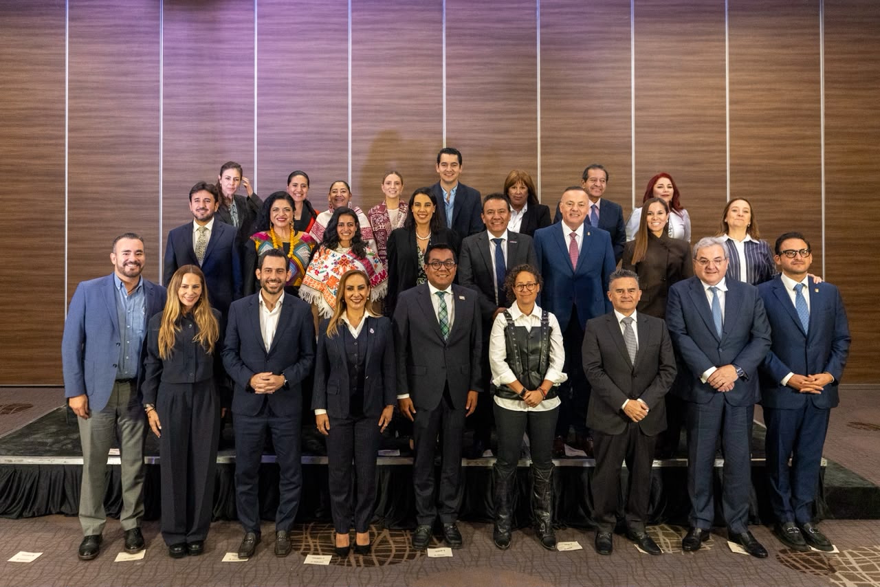 Una Fotografía De Grupo Formal Con Hombres Y Mujeres De Pie En Tres Filas Sobre Una Pequeña Tarima Frente A Una Pared Con Paneles De Madera Rústica En Tonos Marrones Y Morados. La Mayoría De Los Asistentes Visten Trajes Oscuros, Mientras Que Algunas Mujeres Lucen Blusas Tradicionales Mexicanas Bordadas A Mano En Colores Vibrantes. Todos Los Participantes Sonríen A La Cámara En Una Pose Profesional Y Ordenada.