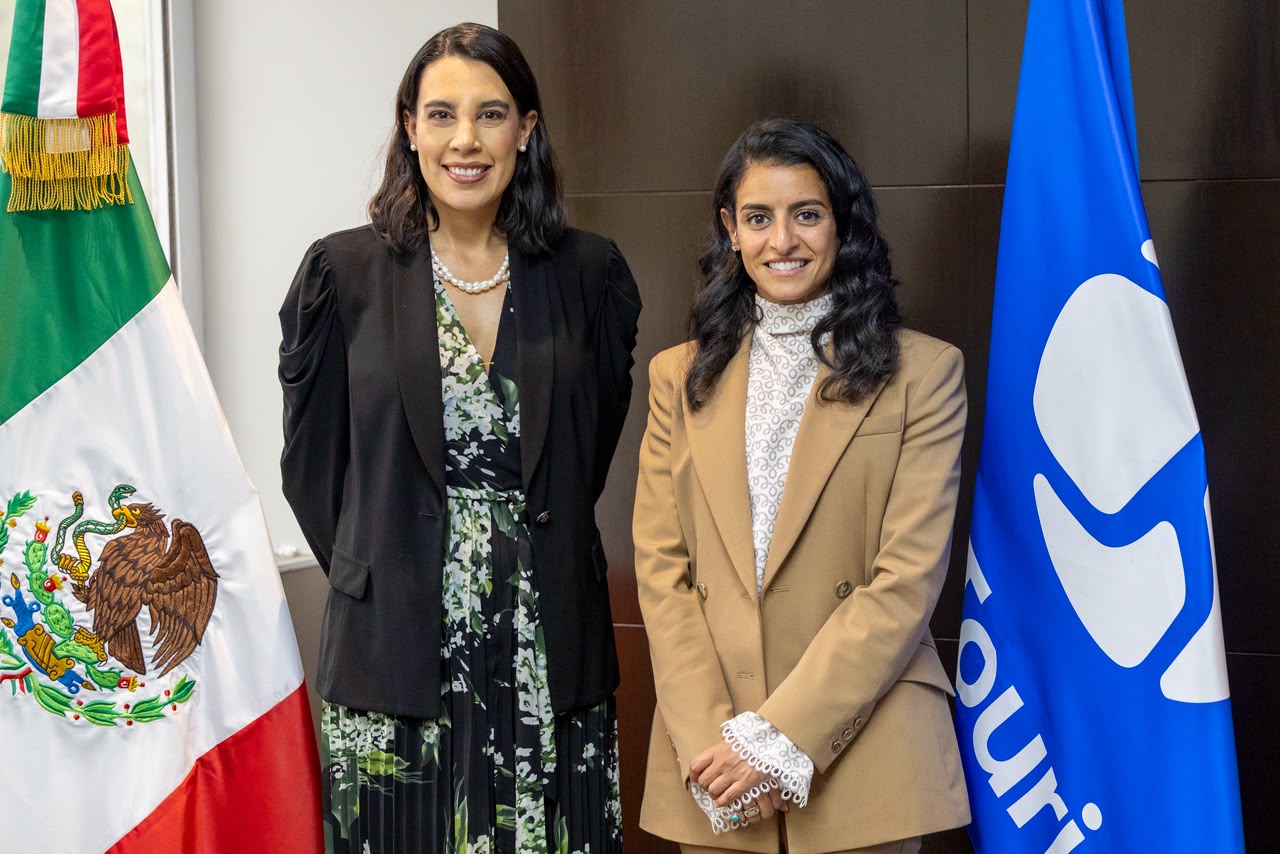 Una Fotografía De Plano Medio De Dos Mujeres Sonrientes De Pie Una Al Lado De La Otra En Una Oficina Formal. A La Izquierda, Una Mujer Con Un Blazer Negro Sobre Un Vestido Floral Y Un Collar De Perlas. A La Derecha, Una Mujer Con Un Blazer Color Camello Sobre Una Blusa Blanca Y Manos Entrelazadas. Están Flanqueadas Por La Bandera De México A La Izquierda Y Una Bandera Azul Corporativa Con Un Logotipo A La Derecha, Frente A Una Pared De Paneles De Madera.
