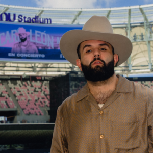 Primer Plano De Medio Cuerpo Del Cantante Carín León, Con Barba Poblada Y Sombrero Beige, Posando En El Campo De Un Estadio Vacío. Al Fondo, Una Pantalla Gigante Lo Anuncia Como &Amp;Quot;Carín León En Concierto&Amp;Quot; En El Ou Stadium.