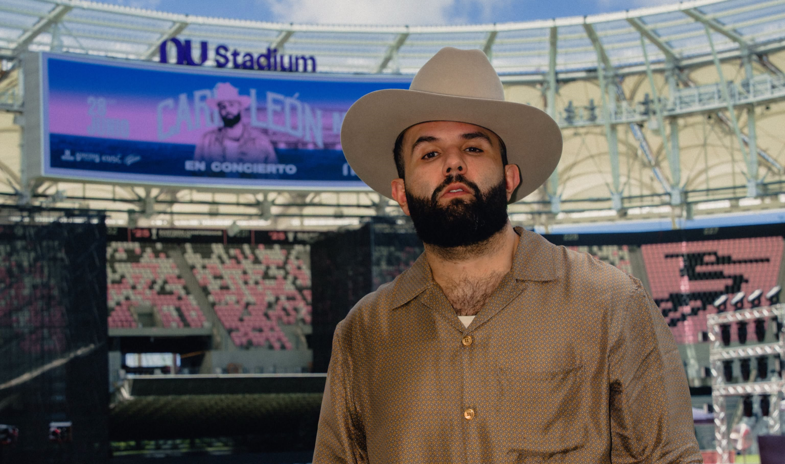 Primer Plano De Medio Cuerpo Del Cantante Carín León, Con Barba Poblada Y Sombrero Beige, Posando En El Campo De Un Estadio Vacío. Al Fondo, Una Pantalla Gigante Lo Anuncia Como &Quot;Carín León En Concierto&Quot; En El Ou Stadium.