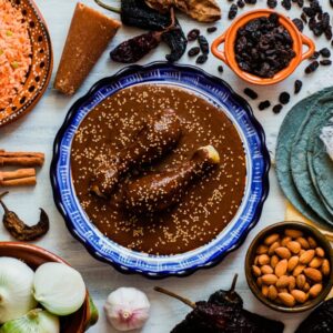 Ruta de los siete moles: un viaje culinario por el alma y la tradición de oaxaca la revista del sureste mx Un Plato Central Con Mole Cubierto De Ajonjolí Y Piezas De Pollo, Rodeado De Ingredientes Como Chiles Secos, Arroz Rojo, Cebollas, Almendras Y Tortillas Azules.