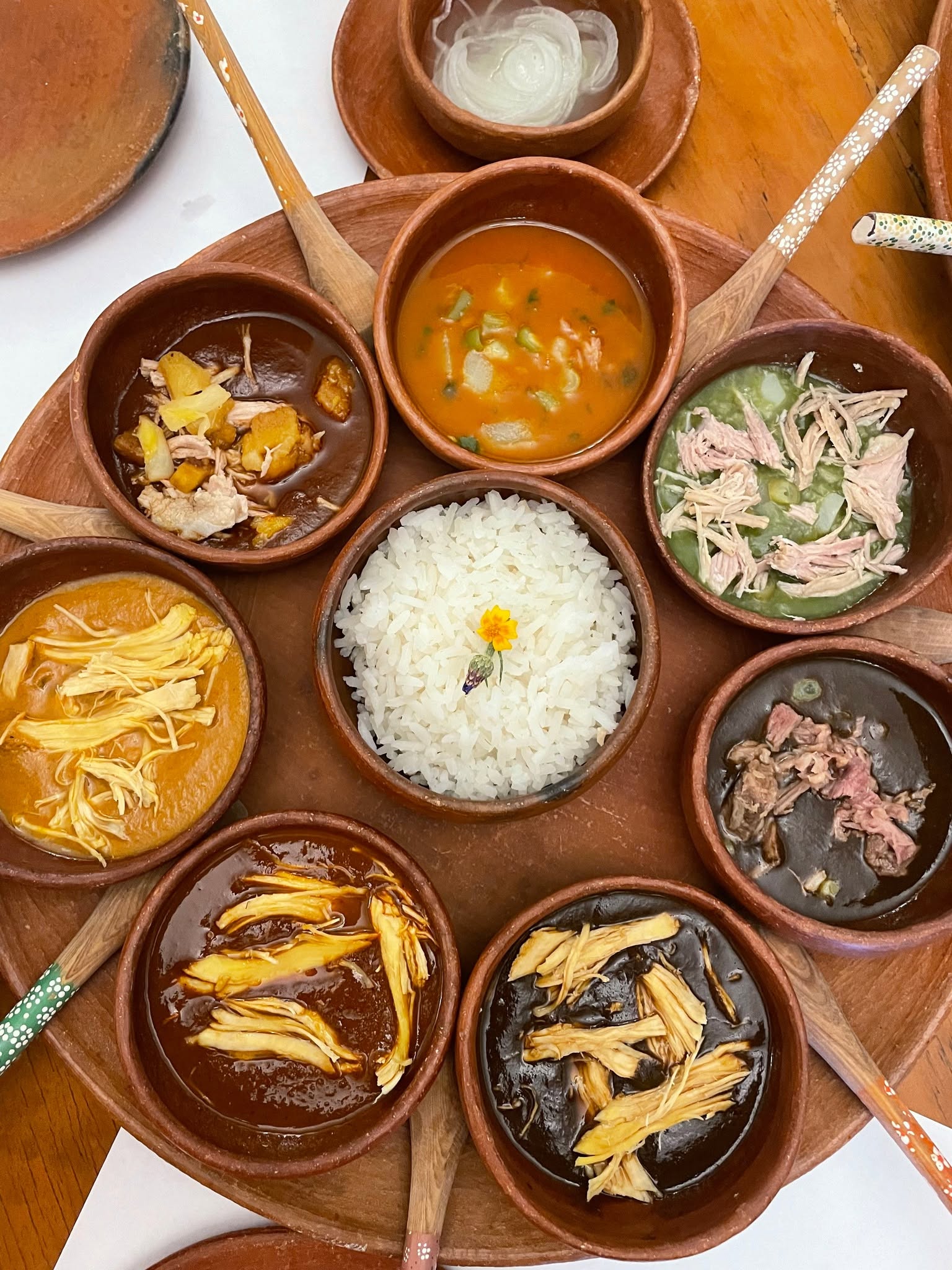 Ruta de los siete moles: un viaje culinario por el alma y la tradición de oaxaca la revista del sureste mx Una Bandeja Circular De Barro Con Varios Cuencos Pequeños Alrededor Que Contienen Diferentes Tipos De Moles Oaxaqueños Y Un Cuenco Central Con Arroz Blanco Adornado Con Una Flor.